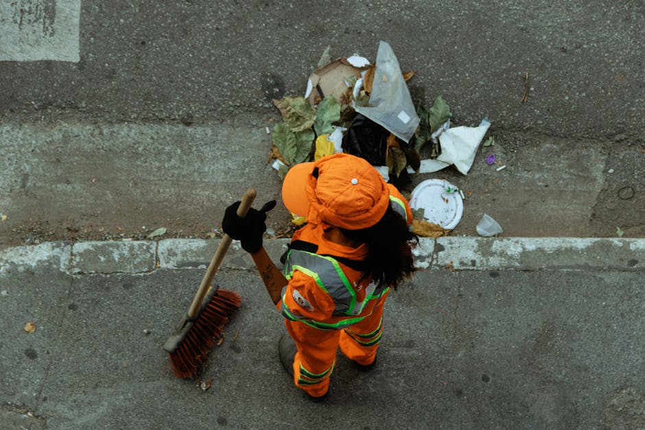 A street cleaner in high-visibility orange and green uniform, wearing a matching orange hat, is sweeping debris and trash on a concrete sidewalk next to a wall with visible stains and dirt. The cleaner holds a wooden-handled broom with red bristles, and the street includes scattered litter such as paper, plastic, and food waste, adjacent to a small patch of an alley or street corridor. The scene is illuminated by natural daylight, emphasizing the cleanup task, as Cleaner Mayfair provides professional sanitation and waste removal services for residential and commercial properties, including bulky waste removal for Mayfair flats and surface cleaning for various urban environments.