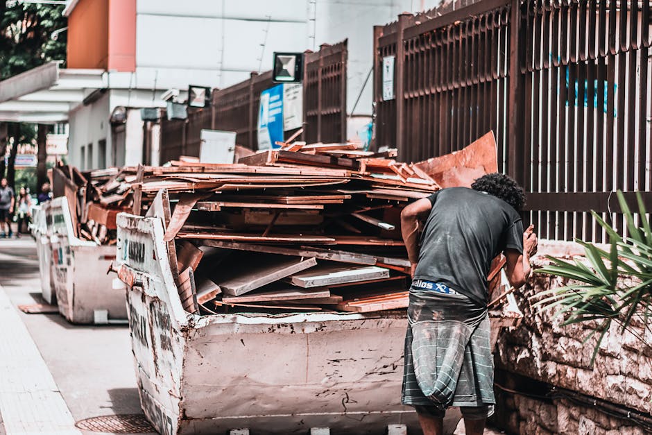 A person with curly black hair wearing a black t-shirt and plaid shorts is standing next to a large, heavily loaded skip filled with discarded wooden panels, tiles, and construction debris. The skip is placed on a pavement beside a tall brown metal fence, which runs along the roadside in Mayfair. In the background, blurred city buildings and pedestrians are visible under bright daylight. This scene illustrates waste removal or clearance process, emphasizing the importance of proper disposal and site cleaning, aligning with Cleaner Mayfair's services for bulky waste removal for Mayfair flats. The area appears clean outside of the waste collection, with no dirt or debris on the pavement itself.