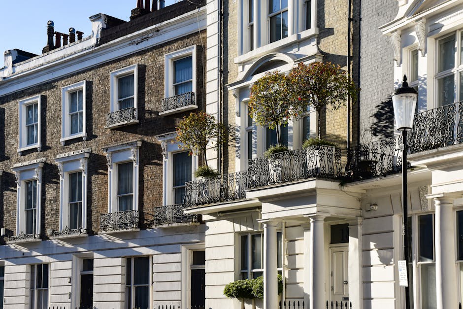 Exterior view of residential homes on Berkeley Square in Mayfair, showing a row of Georgian-style buildings with brick facades and white decorative trim. The buildings feature small balconies with ornate wrought-iron railings, potted plants, and a traditional black street lamp on the sidewalk. The scene is well-lit with natural daylight, highlighting the clean, well-maintained surfaces of the building exteriors. Cleanermayfair.org.uk specializes in surface cleaning, deep cleaning, and sanitisation for such historic homes, ensuring their features remain pristine and hygienic.
