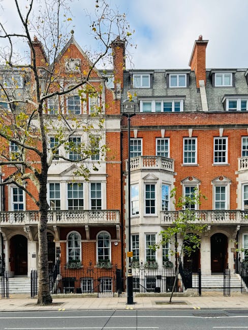 Image of a row of elegant, multi-story residential buildings in Mayfair with red brick facades, white decorative stonework, and large sash windows. The buildings feature small balconies with ornate railings and are set against a partly cloudy sky. In front, a leafless tree with branches extending across the scene and a paved pavement with black iron fencing separates the residences from the street. This urban setting exemplifies the historic and upscale character of Berkeley Square homes, emphasizing the importance of regular domestic cleaning and surface maintenance. Cleaner Mayfair specializes in comprehensive cleaning services, helping to maintain the pristine appearance and hygiene of such distinguished properties.