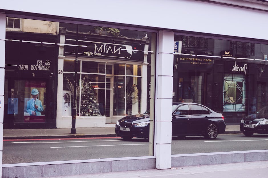A street scene viewed through large glass windows of a commercial building in Mayfair, near Bond Street, showing reflections of the street, parked black and silver cars on the road, and storefronts with signage. Inside, the reflection hints at a clean, well-maintained interior typical of boutique establishments. The glass surfaces appear clean and polished, with no visible smudges or dirt, emphasizing surface cleaning and hygiene maintained by Cleaner Mayfair, specialists in domestic and commercial cleaning near Bond Street.