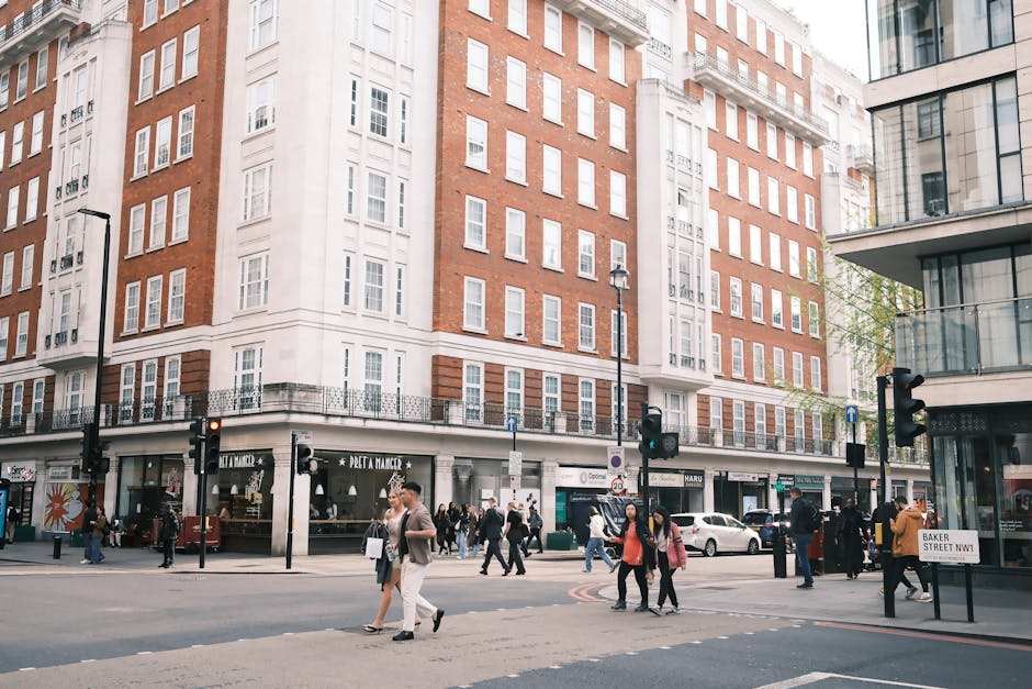 The image depicts a busy street scene in Mayfair near Bond Street, with a large multi-story building featuring a white facade and numerous windows. Ground level shops, including Pret A Manger and other storefronts, are visible with pedestrians walking along the sidewalk and crossing the street at a traffic light. The street is lined with black lampposts and the pavement is clean and well-maintained. Several people are engaged in their daily activities, dressed in casual and business attire. The overall scene shows a vibrant urban environment with modern glass-paneled buildings adjacent to the traditional brick structure, illuminated by daylight. Cleaner Mayfair provides professional domestic cleaning and surface cleaning services in this highly visited area, ensuring hygiene and cleanliness in both commercial and residential properties near Bond Street.
