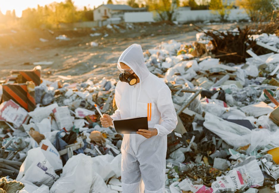 A person wearing a white protective coverall, hood, and goggles is standing in a large outdoor area filled with scattered debris and trash, including plastic bags, cardboard, and discarded materials. 