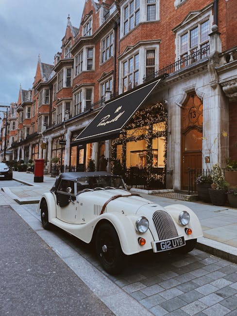 Image depicting a vintage convertible car parked on a cobblestone street in front of an elegant red brick building with large sash windows and decorative stonework, illuminated by soft ambient lighting. The building features a black awning with the word 'Mayfair' and an entrance door with a brass handle. Potted plants decorate the entrance area, enhancing the sophisticated atmosphere of the Mayfair district. This scene captures the historic charm and well-maintained appearance of the area, as seen in the context of residential and commercial streets. The setting shows a clean, well-kept pavement and street, emphasizing cleanliness and urban elegance, aligning with the themes of surface cleaning, deep cleaning, and maintenance as promoted by Cleaner Mayfair in their tips for deep cleaning residential flats on Brook Street and W1K.