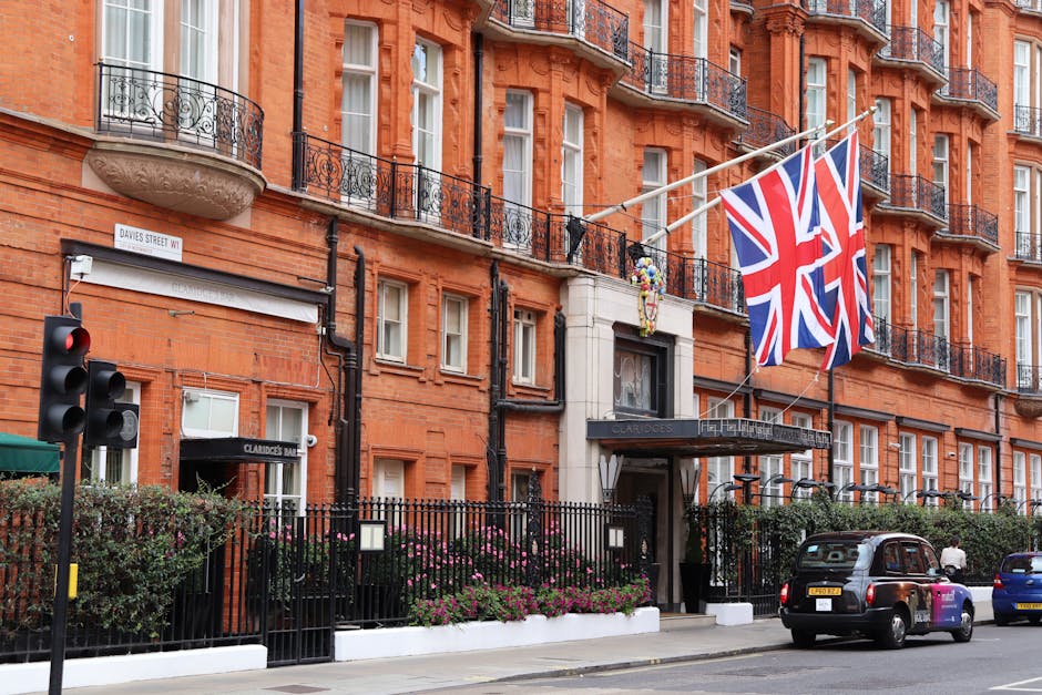 Exterior view of a red brick residential building on Brook Street in Mayfair, featuring multiple windows with white frames and black wrought-iron balconies. The building displays two large Union Jack flags hanging from the facade, with a black iron gate at the entrance adorned with the sign 'Claridge's Bar.' In front, there is a sidewalk with a row of flowering bushes and a traffic light on the street corner. Parked cars, including a black taxi, are visible along the street, and a person can be seen walking nearby. Natural daylight illuminates the scene, highlighting the clean and well-maintained appearance of the building exterior, reflecting high standards of cleanliness associated with professional cleaning services like those offered by Cleaner Mayfair.