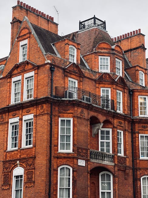 Image depicting a vintage convertible car parked on a cobblestone street in front of an elegant red brick building with large sash windows and decorative stonework, illuminated by soft ambient lighting. The building features a black awning with the word 'Mayfair' and an entrance door with a brass handle. Potted plants decorate the entrance area, enhancing the sophisticated atmosphere of the Mayfair district. This scene captures the historic charm and well-maintained appearance of the area, as seen in the context of residential and commercial streets. The setting shows a clean, well-kept pavement and street, emphasizing cleanliness and urban elegance, aligning with the themes of surface cleaning, deep cleaning, and maintenance as promoted by Cleaner Mayfair in their tips for deep cleaning residential flats on Brook Street and W1K.