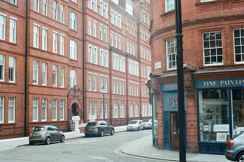 Exterior view of a street in Mayfair W1J, showing red-brick residential buildings with multiple white-framed windows and a corner shop with blue signage, including large glass display windows. Parked cars line the street, which is illuminated by natural daylight, highlighting the cleanliness of the area. The scene depicts a well-maintained urban environment near Grosvenor Square, with no visible litter or dirt, reflecting professional surface cleaning and upkeep, consistent with services offered by Cleaner Mayfair.
