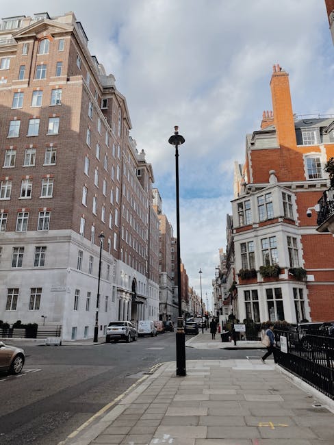 Exterior view of a red-brick residential building on Grosvenor Square in Mayfair, London, with black iron balconies and white-framed windows. Two large Union Jack flags are draped from the facade, and a black iron fence with pink flowers borders the front entrance. A street with parked cars, including a black and a blue vehicle, runs alongside the building, while a traffic light is visible on the corner. The image showcases the historic architecture and vibrant Union Jack decorations typical of this upscale Mayfair area, captured during daylight with natural lighting. Cleaner Mayfair provides expert domestic and commercial cleaning services to maintain the cleanliness and hygiene of such fine properties, ensuring surfaces like windows, floors, and fixtures are thoroughly cleaned and sanitized.