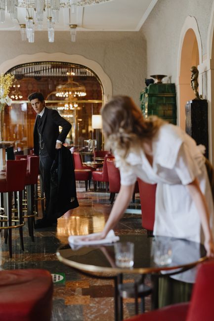 Inside a restaurant with a sophisticated interior, featuring dark wooden floors, red upholstered chairs, and decorative lighting fixtures. A female staff member dressed in a white uniform is cleaning a round wooden table using a cloth, with a glass of water and some small items on the table. In the background, a male staff member dressed in a black suit and apron is standing near another table, with an arched mirror and ornate decorations on the wall behind him. The scene is well-lit, emphasizing the cleanliness and maintained appearance of the space, highlighting surface cleaning and sanitation processes performed by Cleaner Mayfair as part of their restaurant hygiene and deep cleaning services.