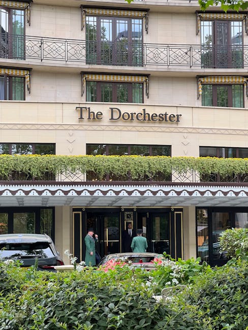 The exterior facade of The Dorchester hotel in Mayfair features large glass doors surrounded by black and gold decorative trims, with a green hedge and lush shrubbery in the foreground. The building has a beige stone finish with ornate iron balconies and large windows. A group of people, dressed in formal attire, are standing near the entrance, which is shaded by a decorative awning. The scene is well-lit, indicating daylight, and the overall appearance emphasizes cleanliness and elegance, consistent with the hotel’s grandeur. Cleaner Mayfair specializes in surface cleaning, deep cleaning, and sanitisation for high-end hospitality establishments like this, ensuring pristine hygiene standards for their customers.