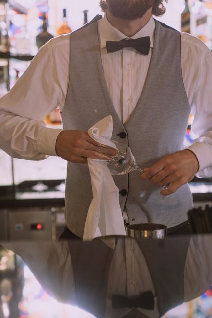 Inside a restaurant with a sophisticated interior, featuring dark wooden floors, red upholstered chairs, and decorative lighting fixtures. A female staff member dressed in a white uniform is cleaning a round wooden table using a cloth, with a glass of water and some small items on the table. In the background, a male staff member dressed in a black suit and apron is standing near another table, with an arched mirror and ornate decorations on the wall behind him. The scene is well-lit, emphasizing the cleanliness and maintained appearance of the space, highlighting surface cleaning and sanitation processes performed by Cleaner Mayfair as part of their restaurant hygiene and deep cleaning services.