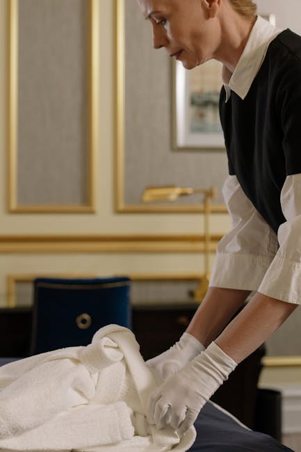 A professional cleaner from Cleaner Mayfair is performing a deep surface cleaning in an elegantly decorated room. She is wearing white gloves and a black and white uniform, carefully folding or arranging white linen on a table covered with a soft, plush white cloth. The background features gold-framed mirrors and muted-colored walls, indicating a luxurious environment. The scene emphasizes meticulous hygiene and thorough cleaning processes, highlighting the company's capability in delivering thorough end-of-lease or domestic cleaning services that restore surfaces, linens, and furniture to a pristine condition. Visible surfaces include the linen, with attention to detail in handling textiles, reflecting a focus on sanitisation and maintenance of high-end residential spaces, suitable for same-day service requests as promoted on the page.